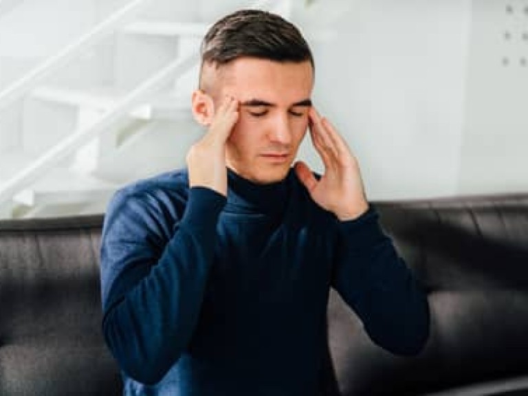 Young handsome man keeps his hands on temples, feeling headache, with closed eyes. Sitting on sofa at home.