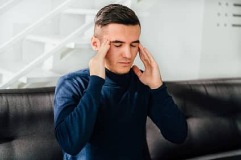 Young handsome man keeps his hands on temples, feeling headache, with closed eyes. Sitting on sofa at home.