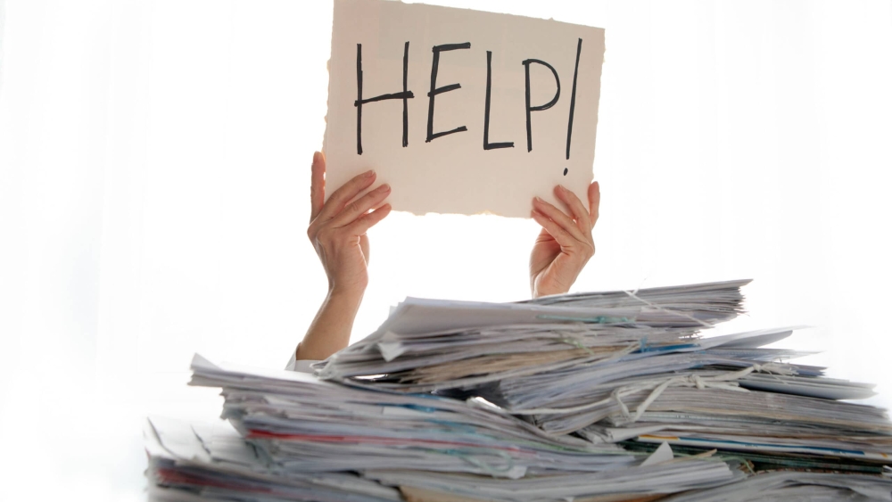 Person under a pile of papers with a hand holding a sign of help. Accounting.