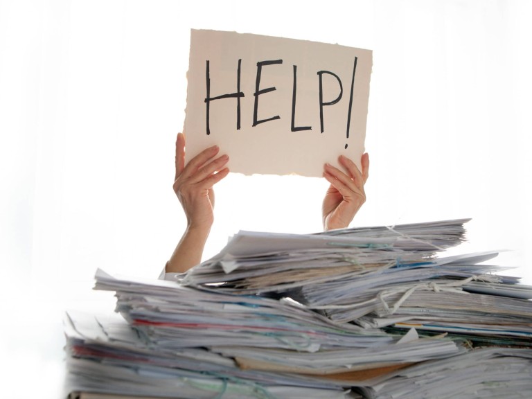 Person under a pile of papers with a hand holding a sign of help. Accounting.